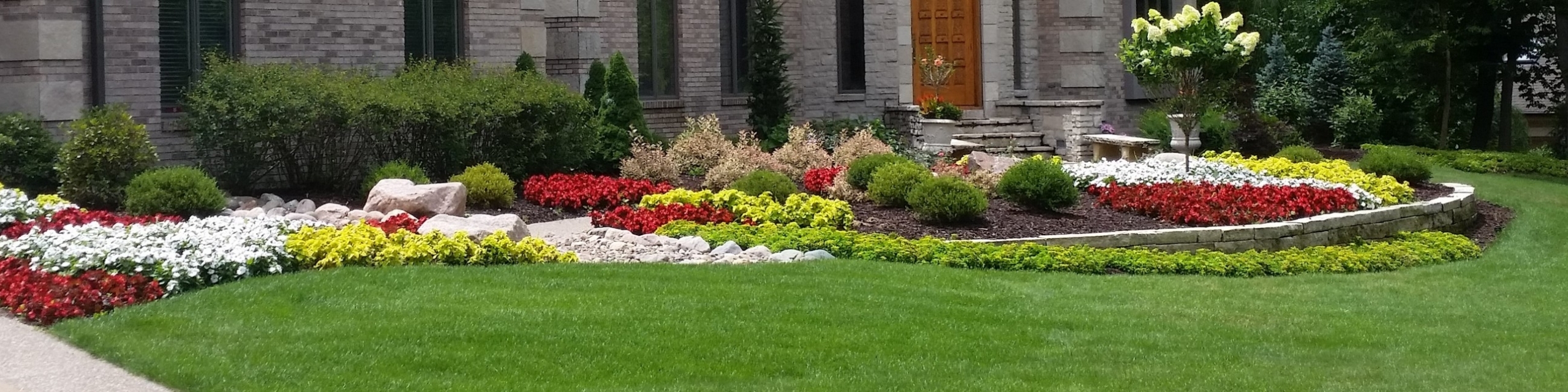 The landscaping in front of a two story home, alongside the home's garage. The red flowers and green bushes dot alongside the concrete walkway to the entrance of the home.