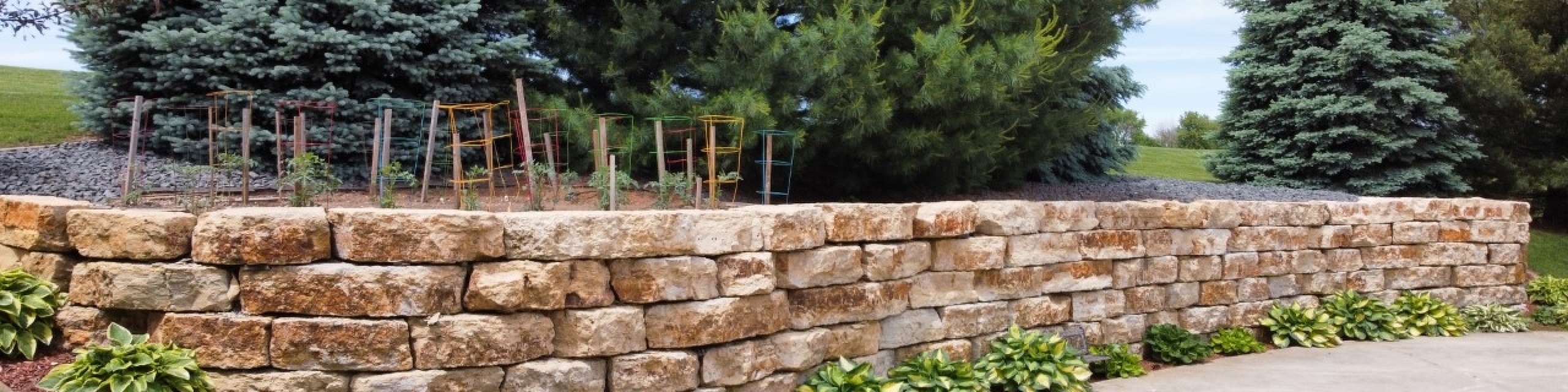 Brick boulder retaining wall made with tan bricks and surrounded by dark red mulch with plants planted in regular intervals.